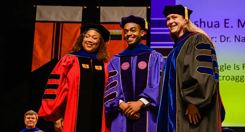 A male doctoral graduate wearing purple academic regalia poses with two female professors — one in red regalia and the other in gray regalia — during the doctoral ceremony.